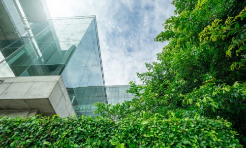 A sky from the angle of looking up at a building, it also features green trees and the corner of a glass building