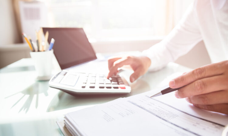 Image shows a close up of a male's hand holding a pen to paper and another hand on a calculator.