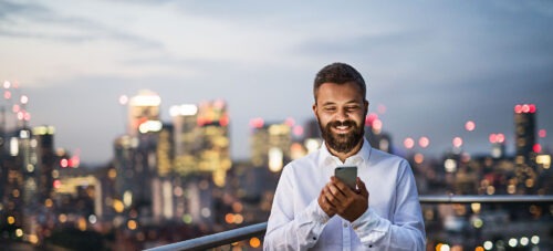 A man standing on a rooftop with a blurred skyline in the background looking at the screen of his phone.