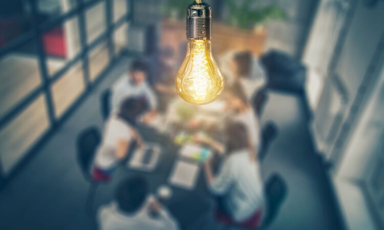 A birds eye view angle showing a lit up bulb in the centre of a ceiling with a blurred view of office workers around a table below.