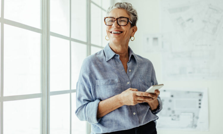 A white female with short grey hair standing in front of a panelled window with her phone in her hand, smiling.