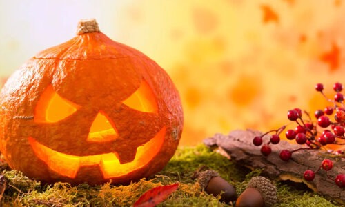 A pumpkin with cut out eyes and mouth against a warm orange back drop on moss.