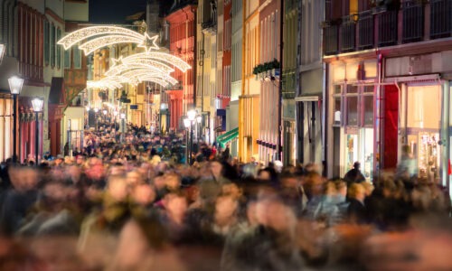 A busy high street during the Christmas shopping period. The people are blurred out and you can see shops and Christmas lights in the distance.