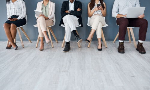 Five people sitting down on chairs, only their legs and feet are seen. It is like lining up at a job interview.