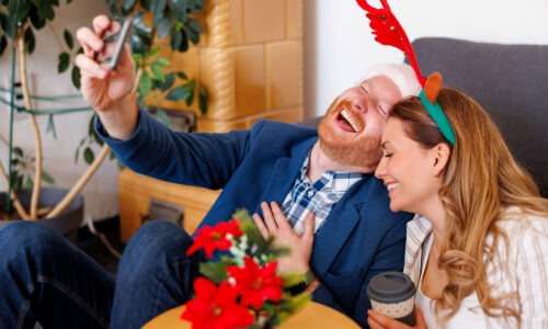 A white male and female taking a selfie with their phone and laughing while wearing christmas themed hats.
