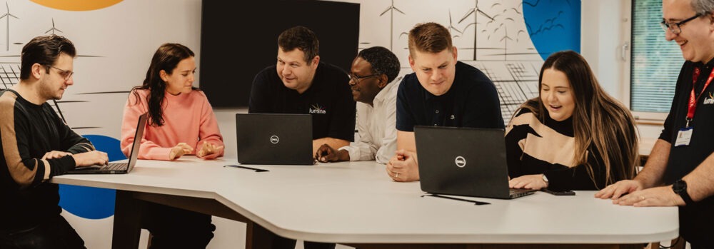 A group of team members from Lumina Energy in the office, at a large table looking at laptops, smiling.